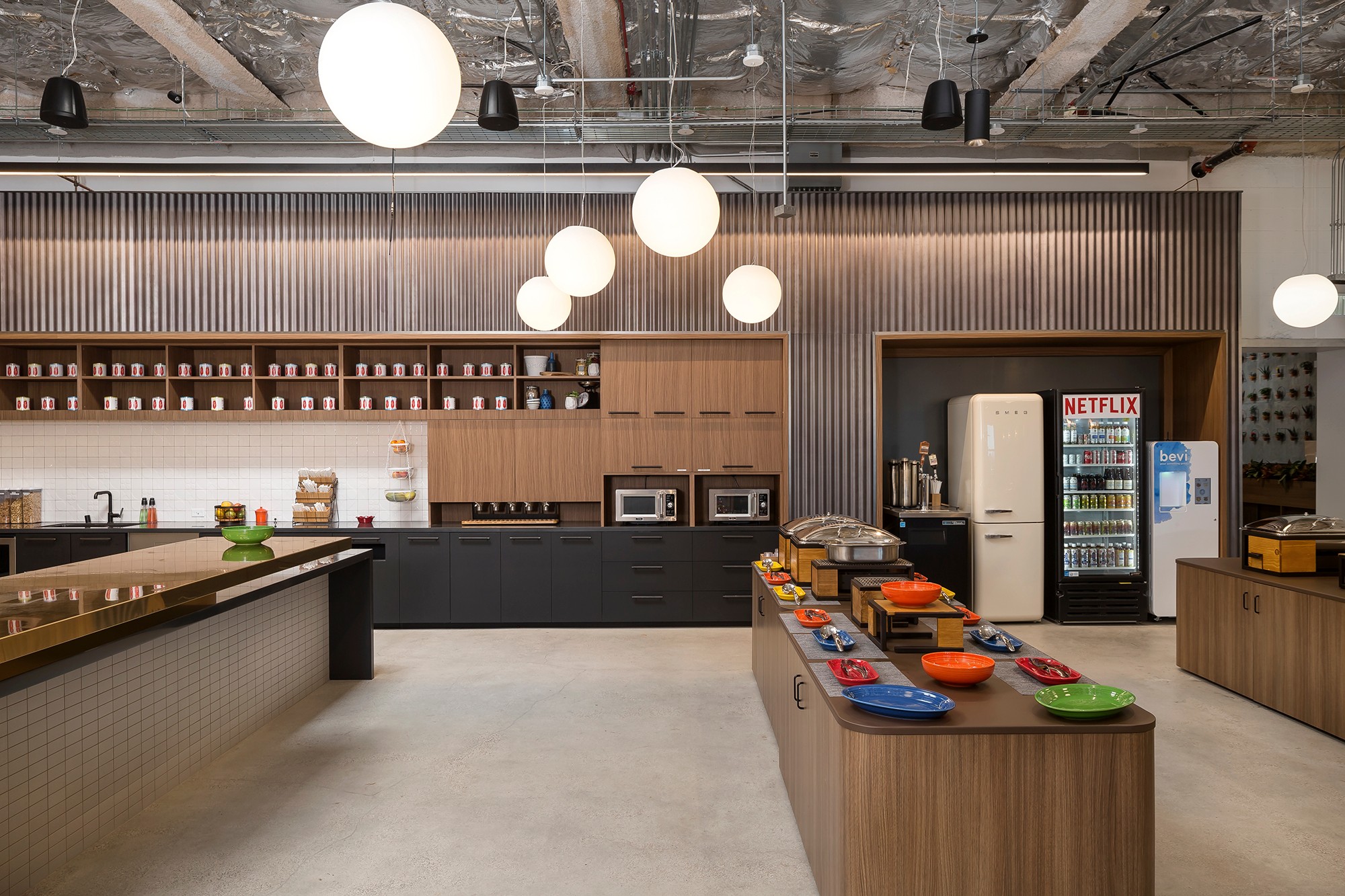 Modern kitchen with hanging lights, wooden shelves, microwave, refrigerator, and a table with colorful bowls.