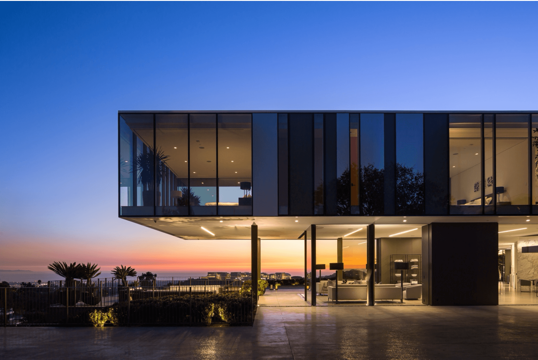 Modern building with glass facade against a sunset sky, with outdoor area and plants in foreground.
