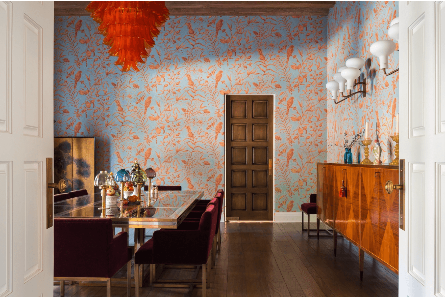 Dining room with patterned wallpaper, wooden door, sideboard, and a table with chairs, decorated with vases and artwork.