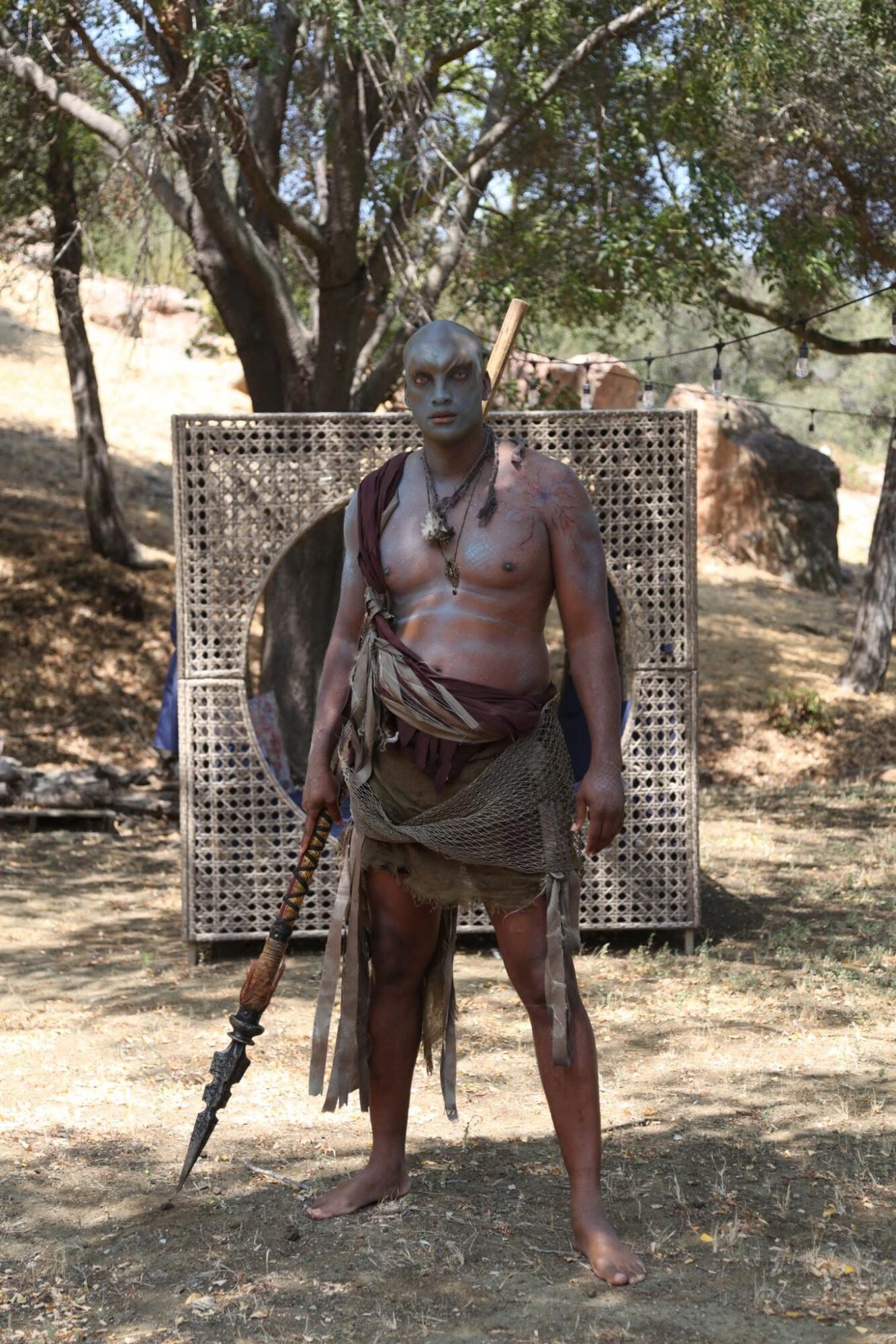 Man standing outdoors with a spear, wearing traditional clothing, trees and rocks in background.