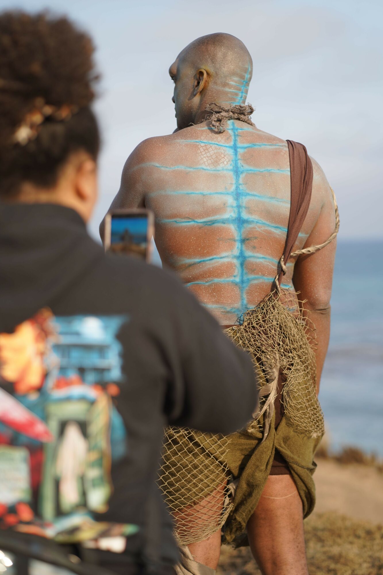 Person with painted blue lines on back, holding a phone, outdoors, with another person nearby, landscape background.