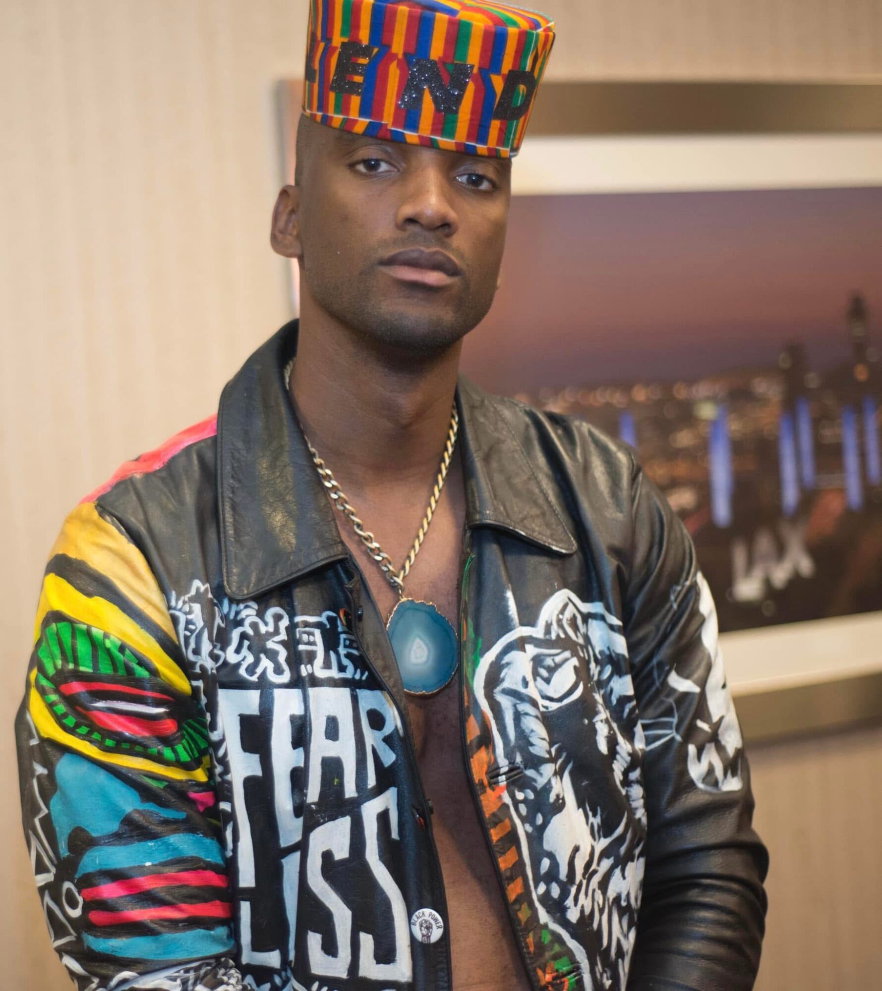 Man wearing a colorful hat and a black jacket with vibrant patterns, standing indoors with a blurred background.