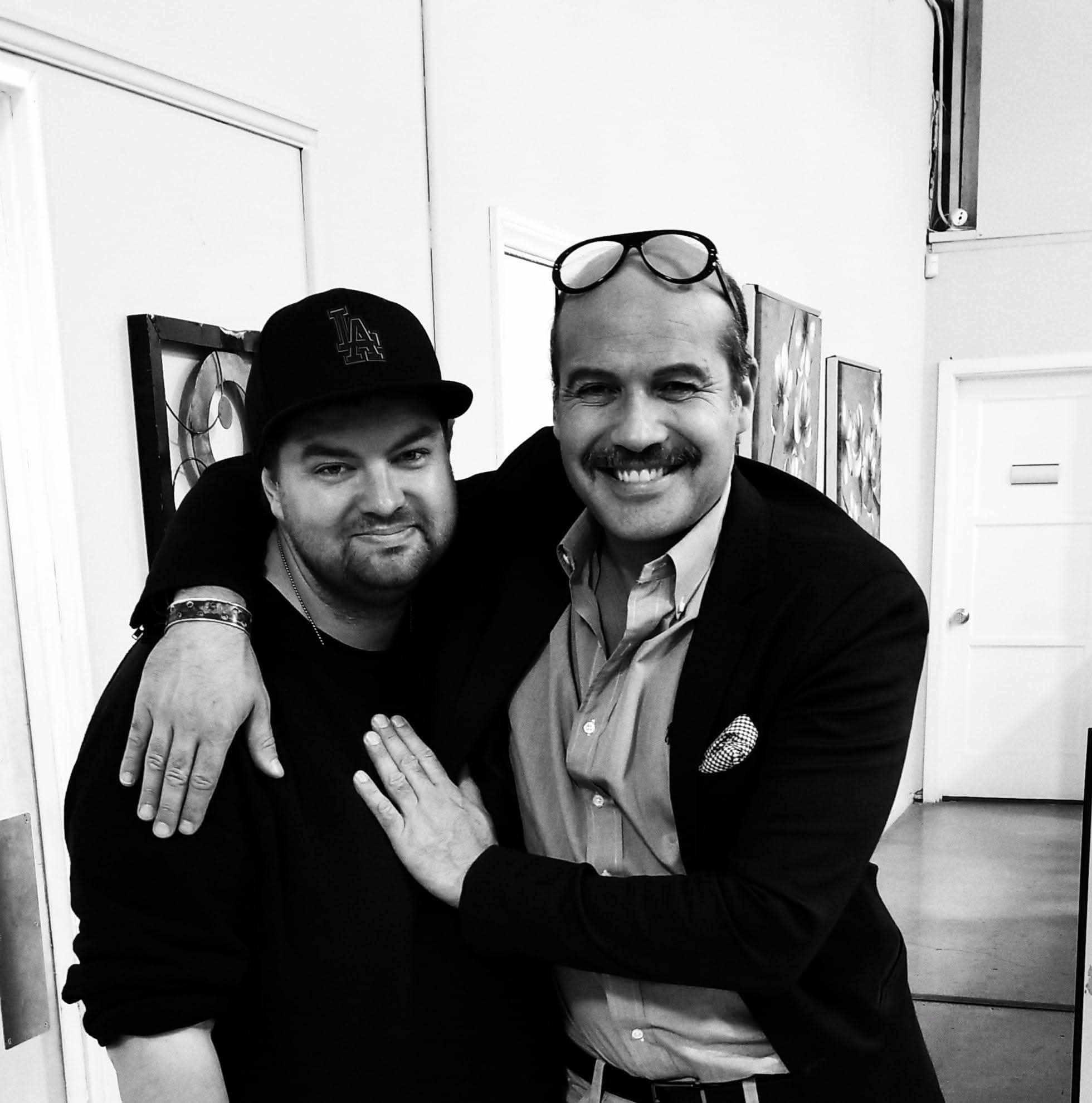 Two men smiling, one with glasses on head, arm around each other, in an indoor setting with artwork on the wall.