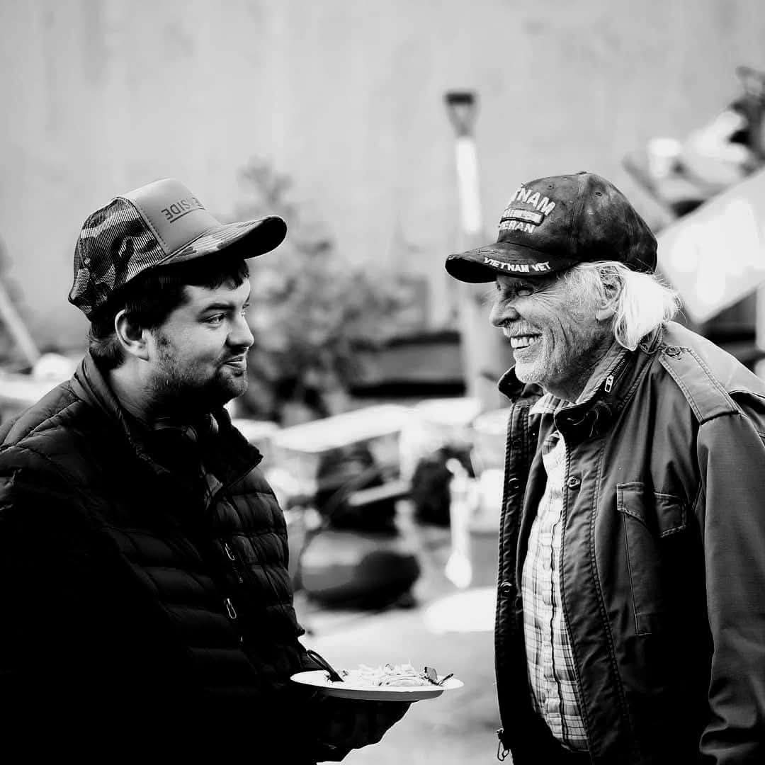 Two men smiling and talking outdoors, one holding a plate, in black and white.
