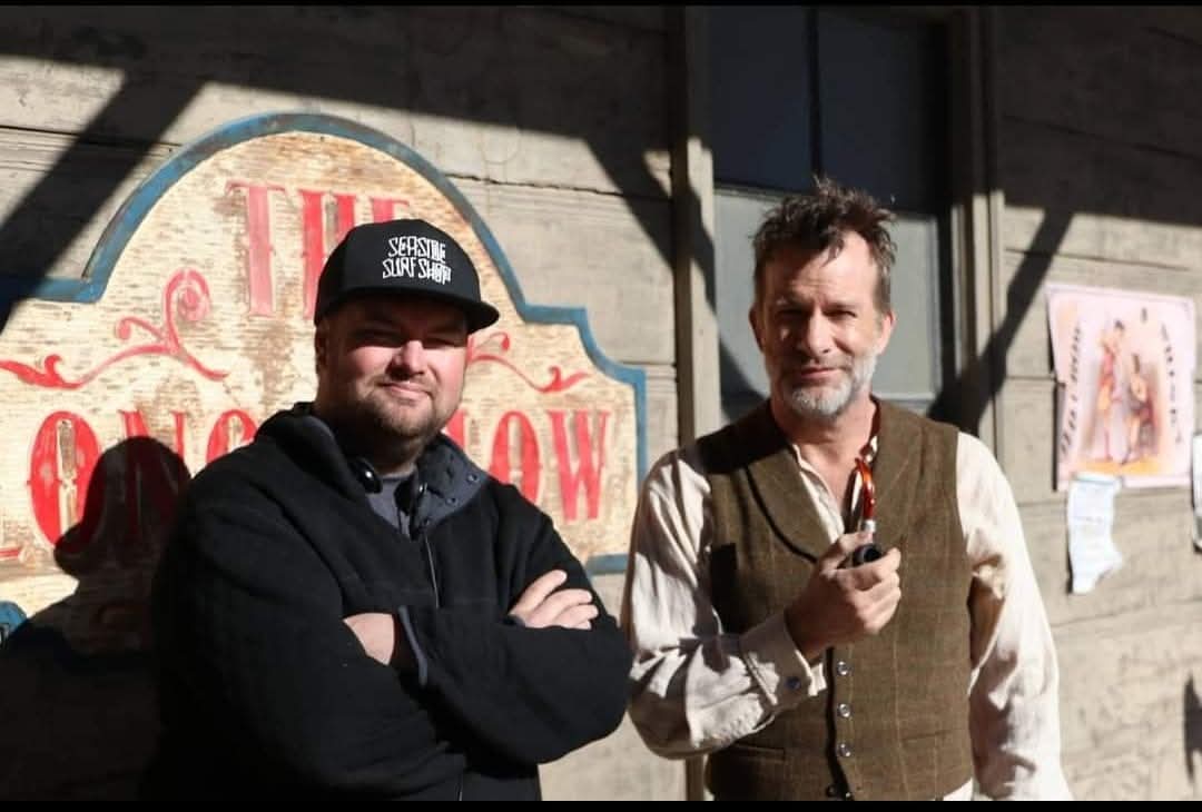 Two men standing outside in sunlight, one with arms crossed and the other holding a pipe, in front of a building with a sign.