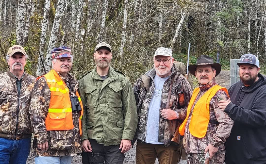 Six people outdoors in a wooded area, some wearing camouflage and safety vests, standing together and smiling.