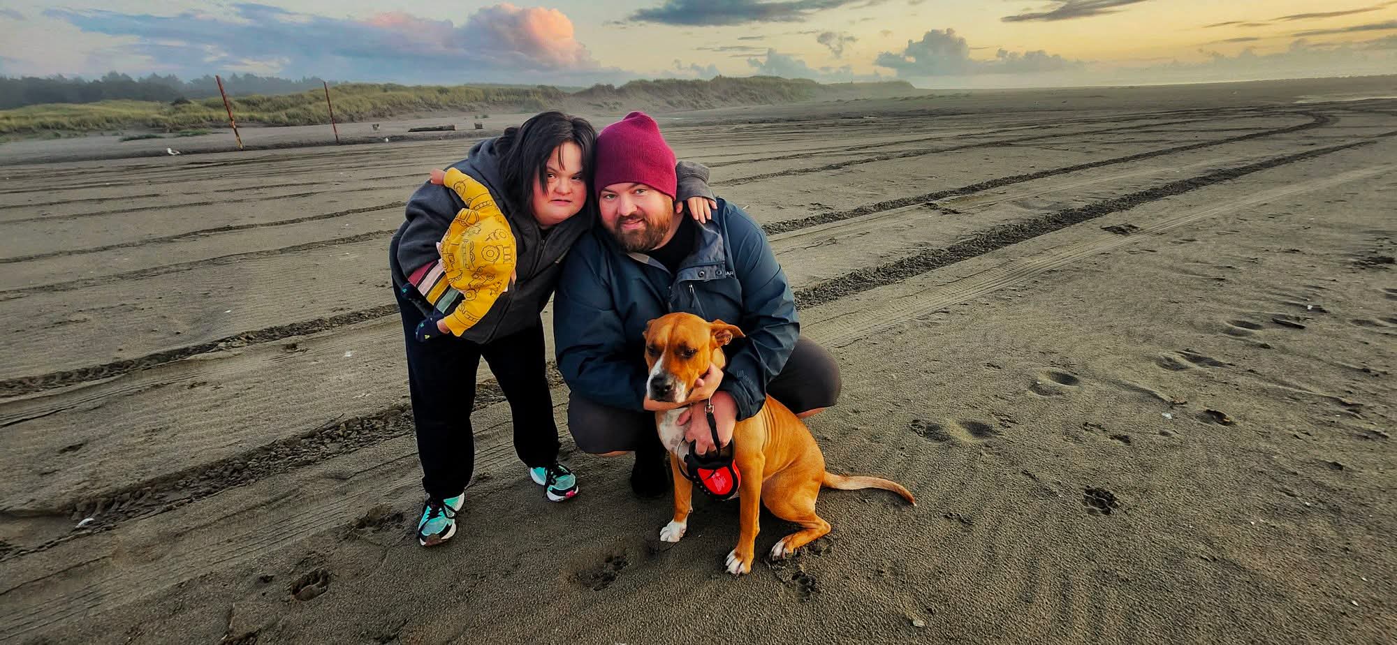 Two children and a dog on a sandy beach during sunset, with the children smiling and the dog sitting calmly.