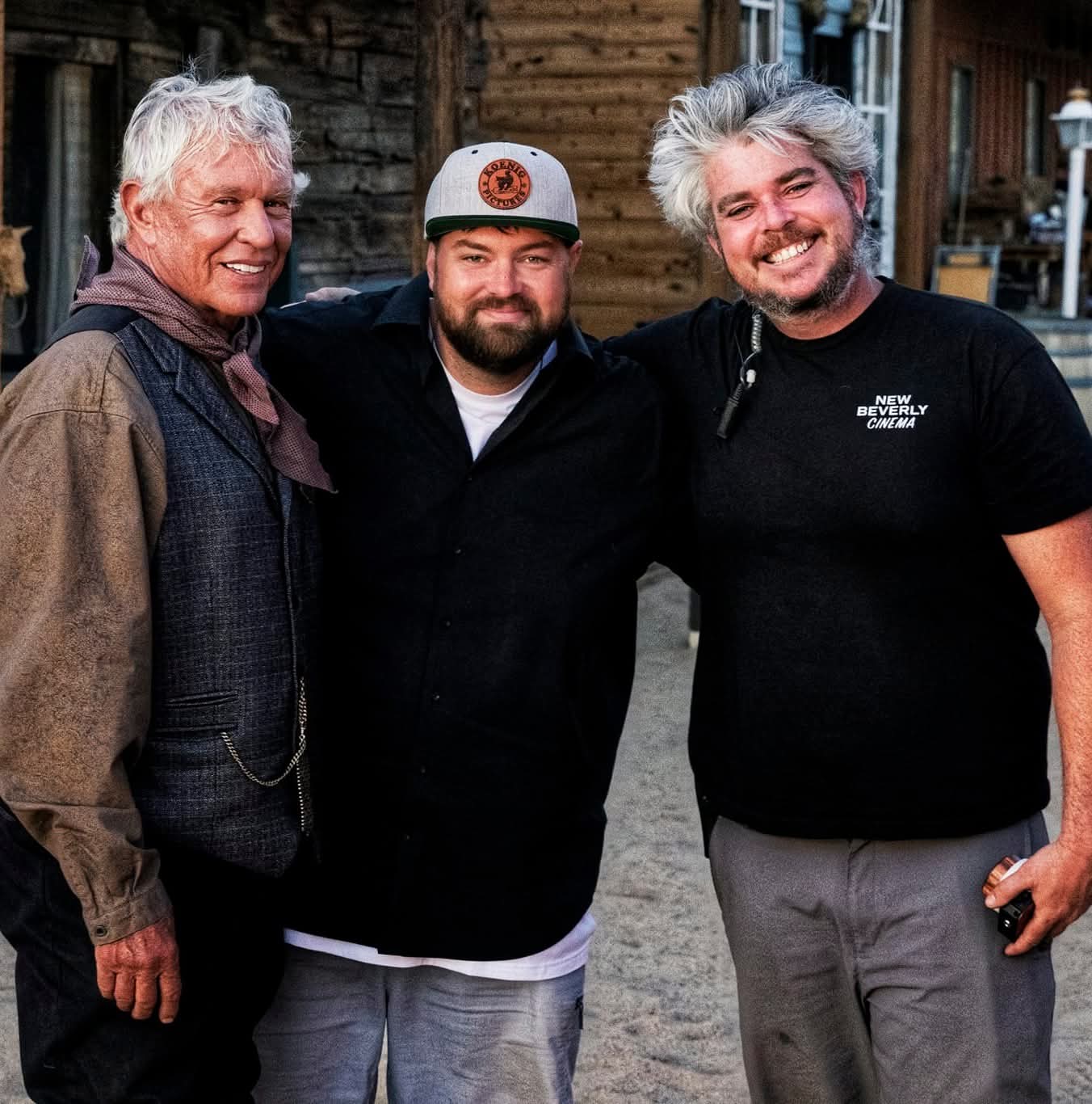Three men smiling outdoors, standing close together, with wooden buildings in the background.