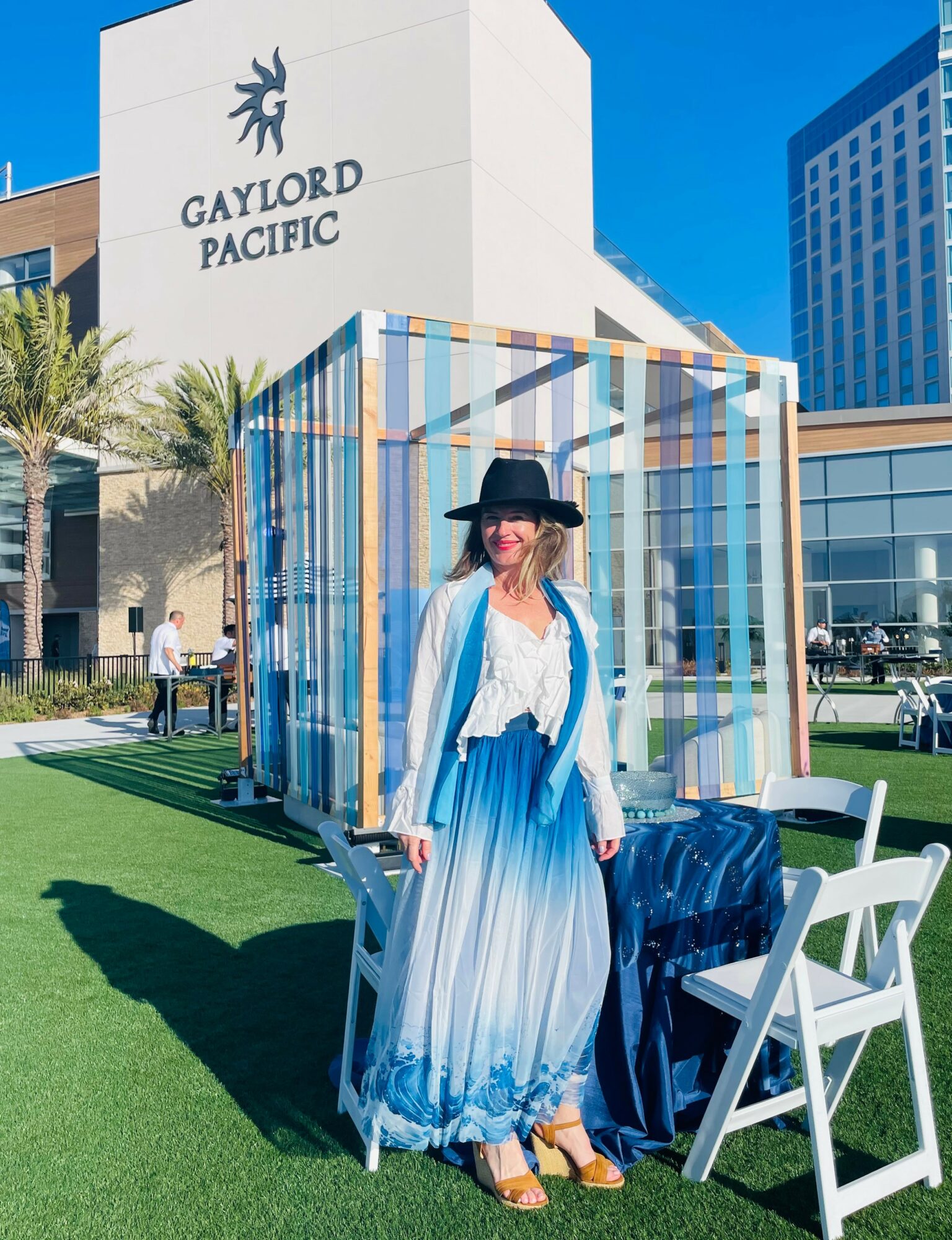 Woman in a hat and blue dress standing outdoors near chairs and a colorful structure, with a building labeled 'Gaylord Pacific' in the background.