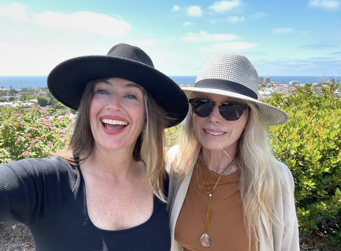 Two women outdoors smiling, wearing hats and sunglasses, with greenery and a blue sky in the background.