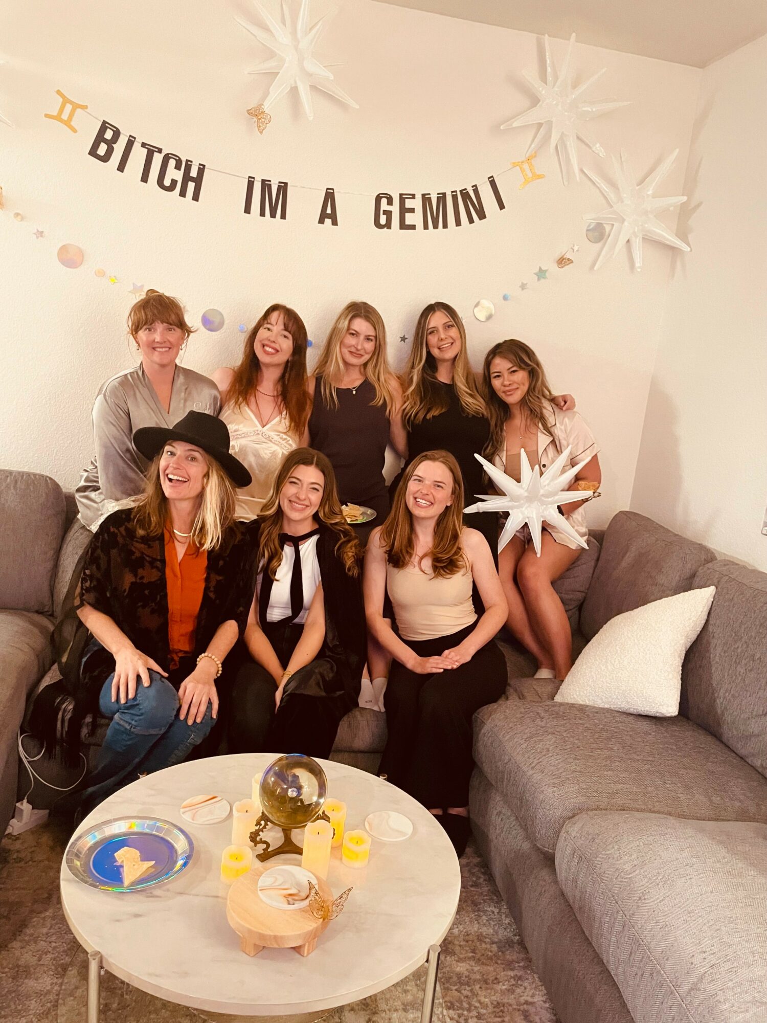 Group of eight women sitting and standing on a sofa, celebrating with decorations and a table of snacks, in a room with star-shaped decorations and a banner reading