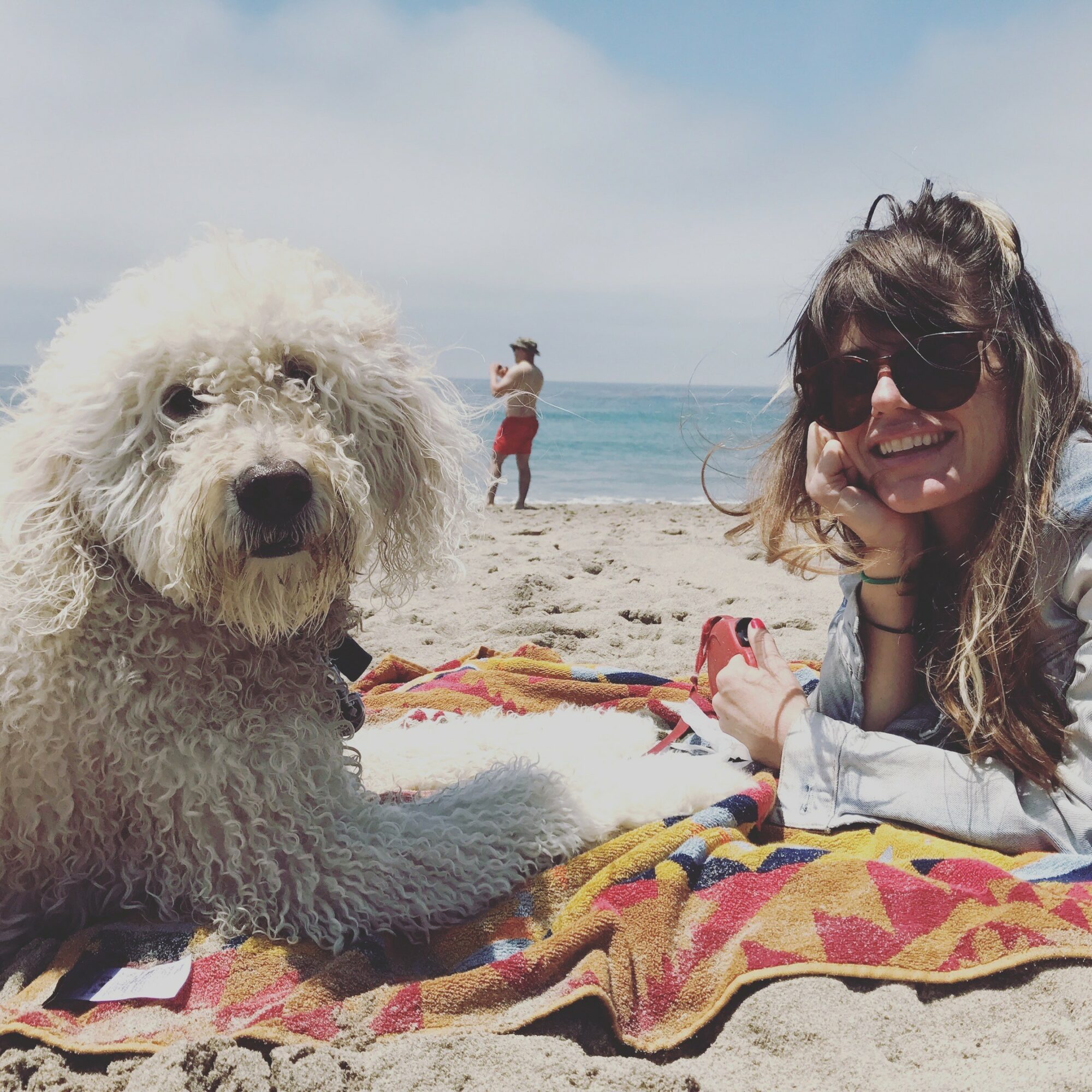 Person with long hair and sunglasses lying on a colorful towel at the beach with a fluffy white dog, ocean in background.