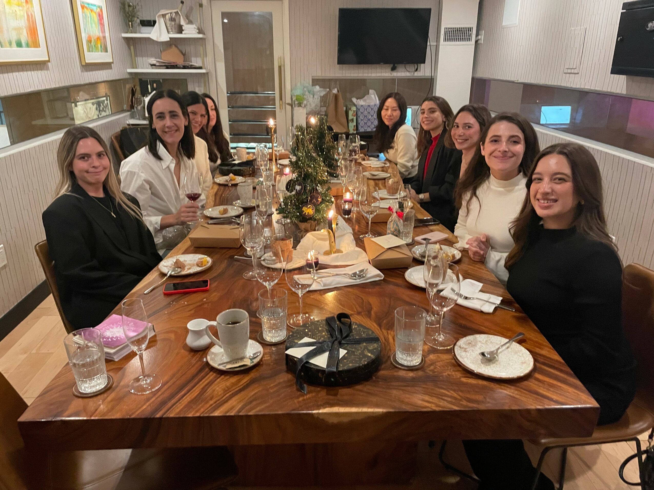Group of ten women sitting around a decorated dining table with food and drinks, in a modern room with a TV and shelves.