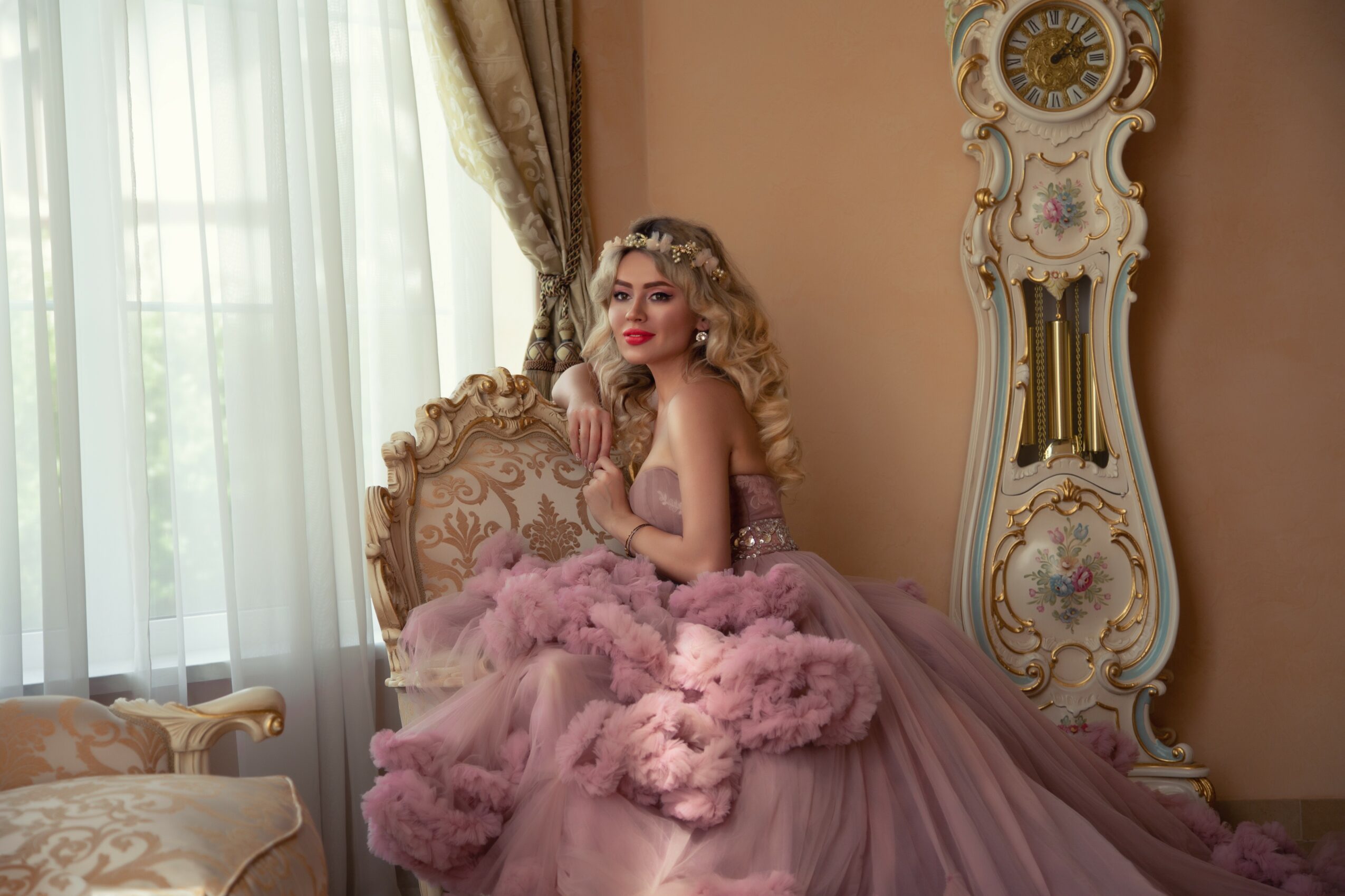 Young woman with curly blonde hair in a pink gown sitting on an ornate chair in a decorated room.