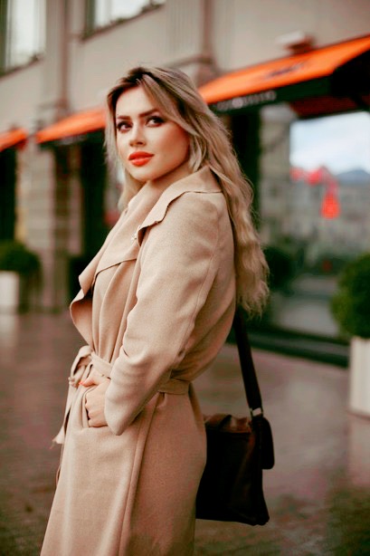 Woman with long hair in beige coat standing outdoors, looking at camera, with blurred background of buildings and greenery.