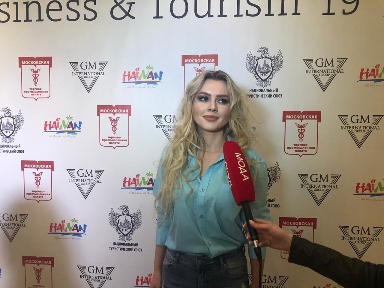 Young woman with long blonde hair being interviewed with a red microphone, standing in front of a patterned backdrop.