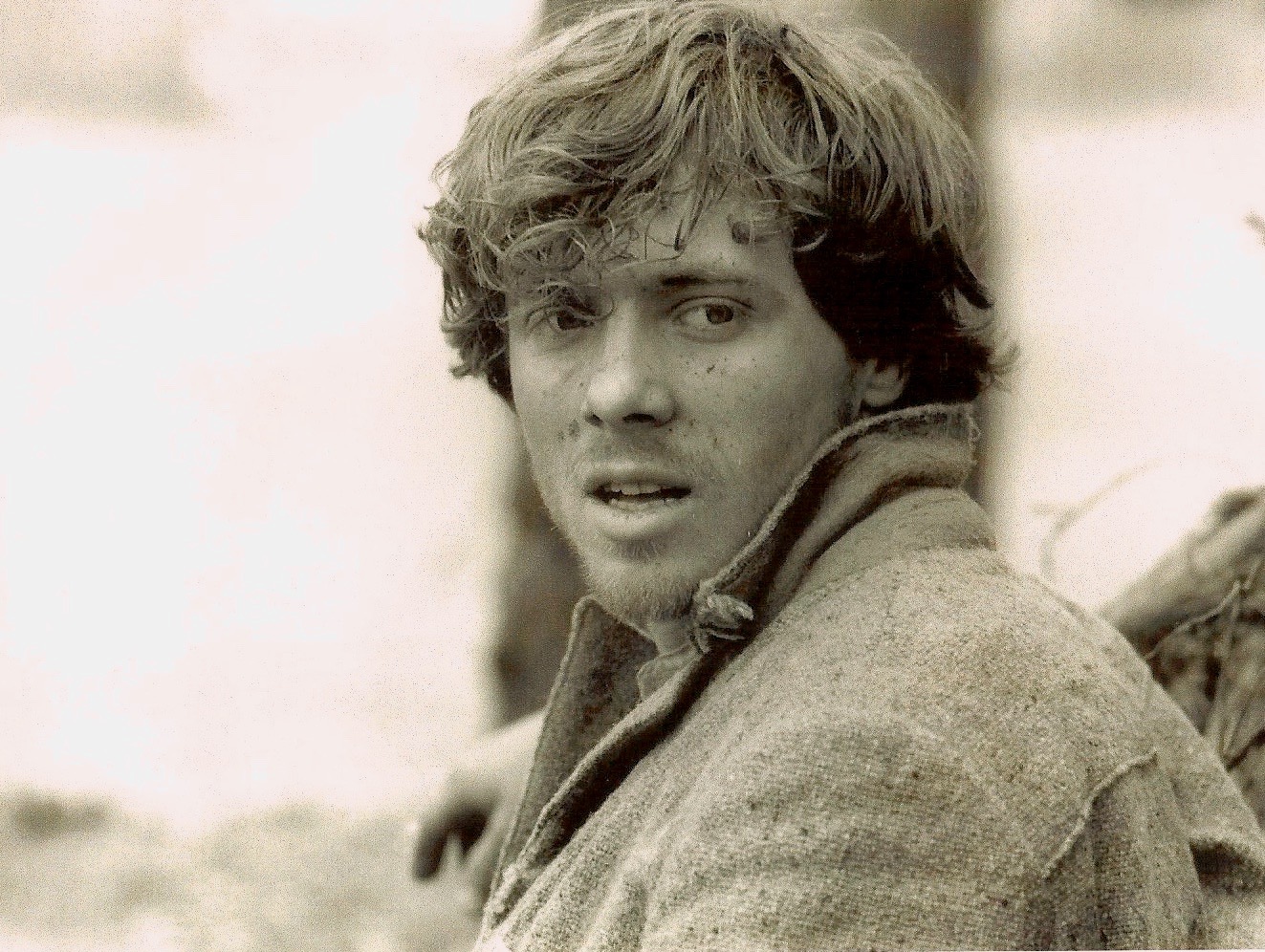 Young man with wavy hair looking at the camera, wearing a coat outdoors, with a blurred background.