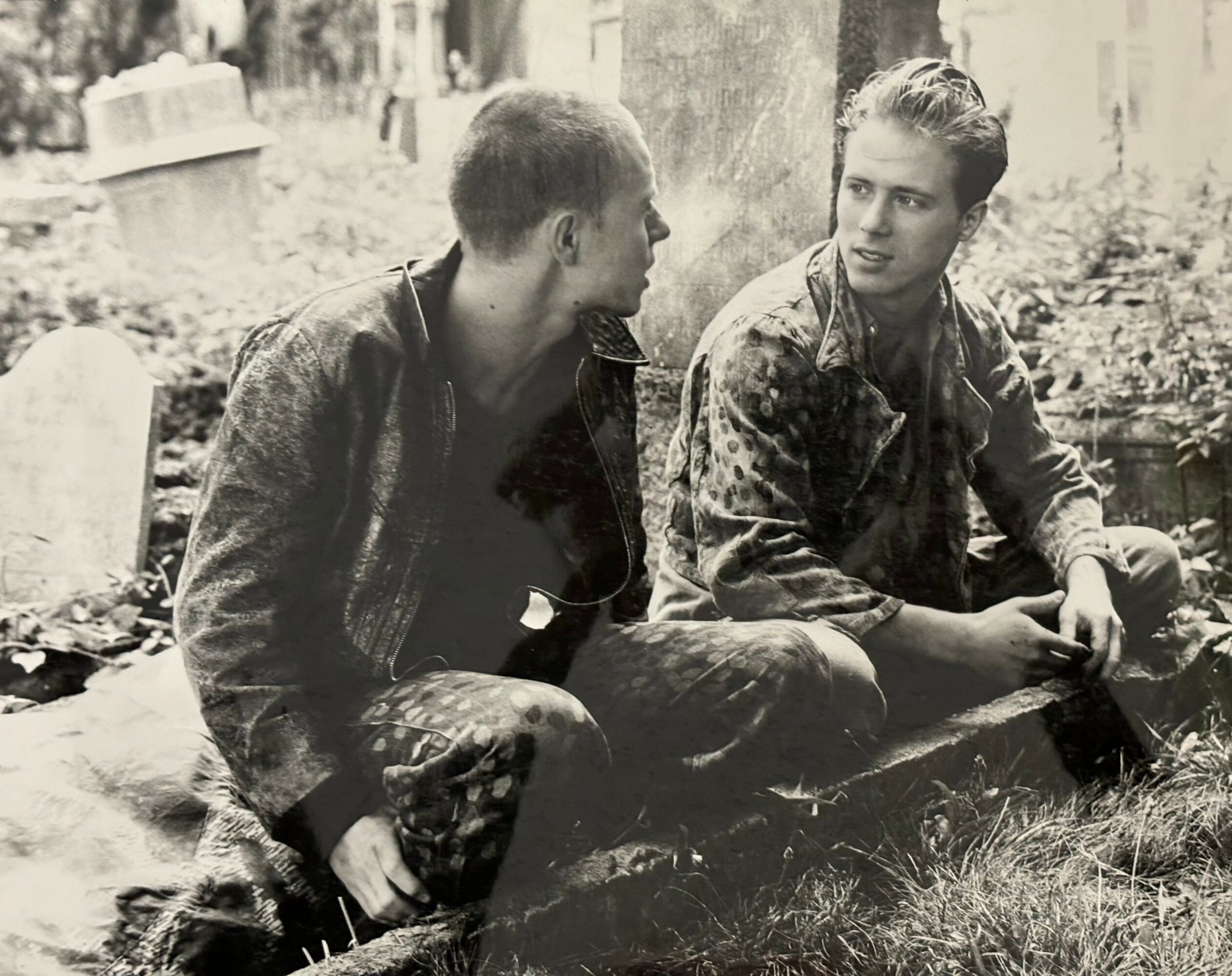 Two young men sit on the ground outdoors, engaged in conversation, with trees and a grave in the background.