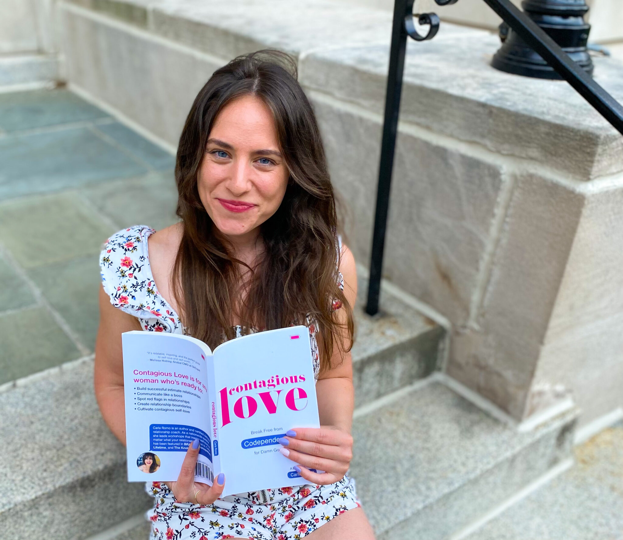 Young woman with long brown hair sitting on stone steps, reading a book outdoors, smiling, wearing a floral dress.