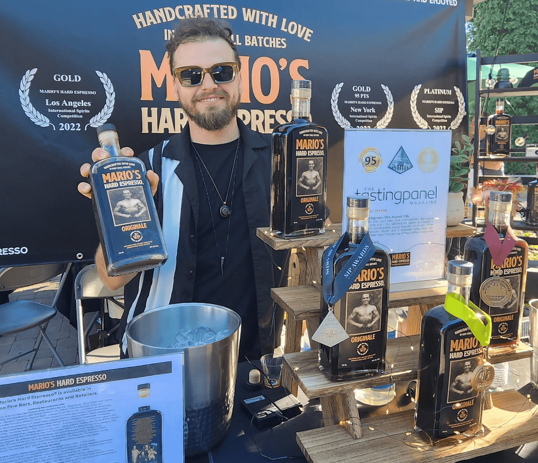 Man with sunglasses and beard holding a coffee box at a booth with coffee products and awards, outdoor setting.