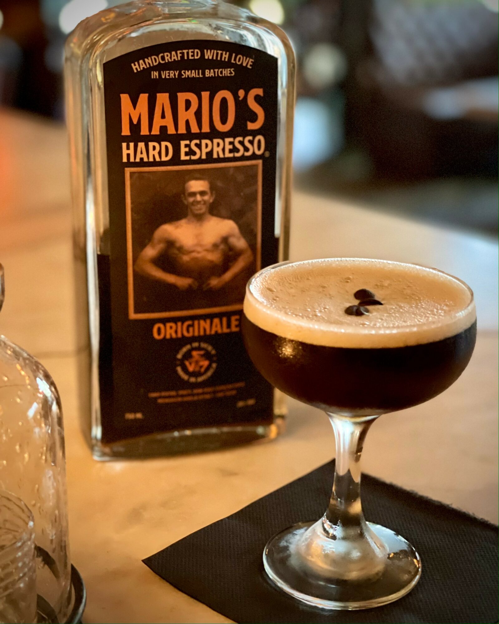 A glass of dark espresso with a foamy top on a black napkin, with a Mario's coffee bottle in the background.