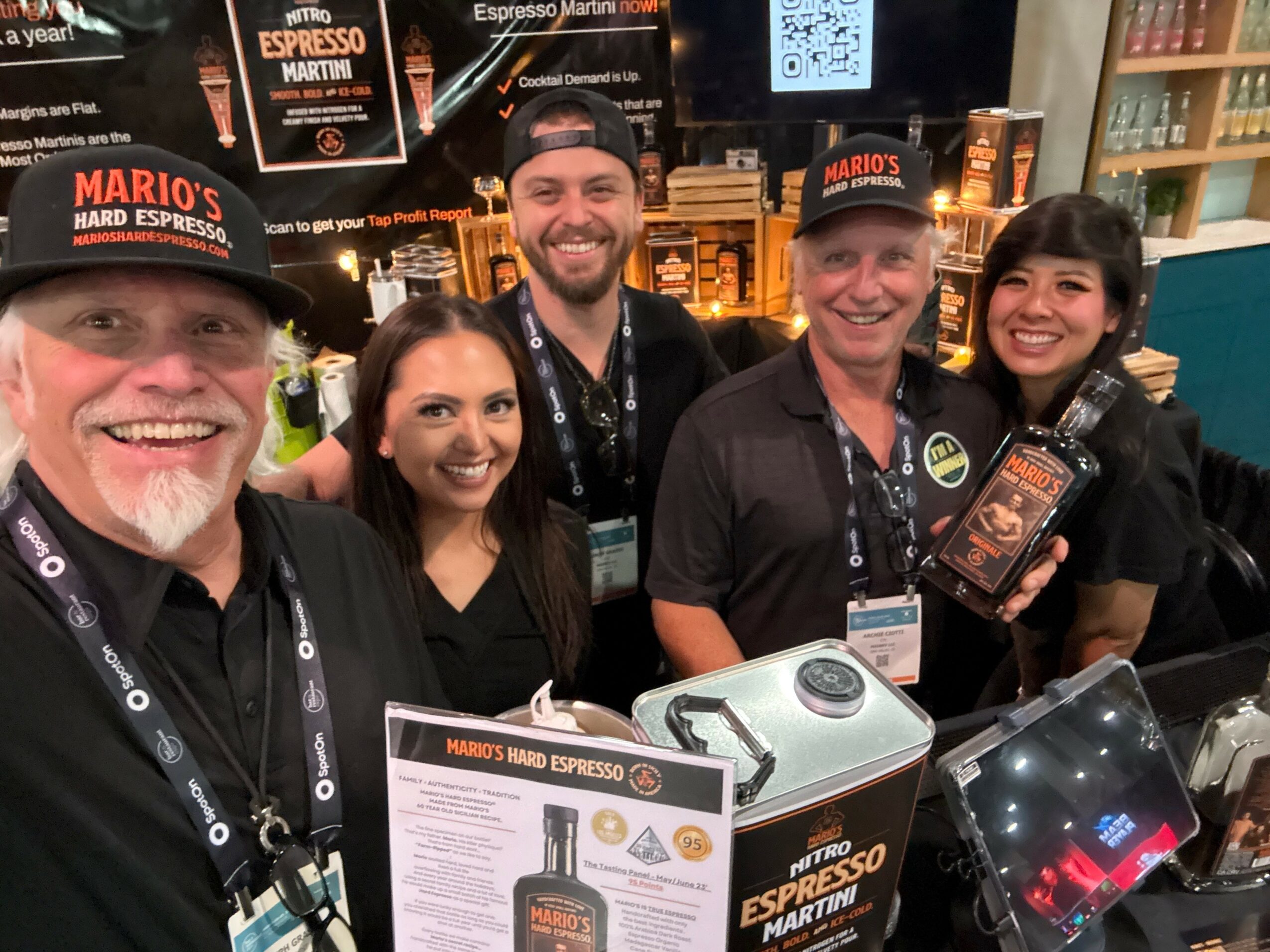 Group of five people smiling behind a coffee shop counter, with coffee equipment and promotional materials visible.