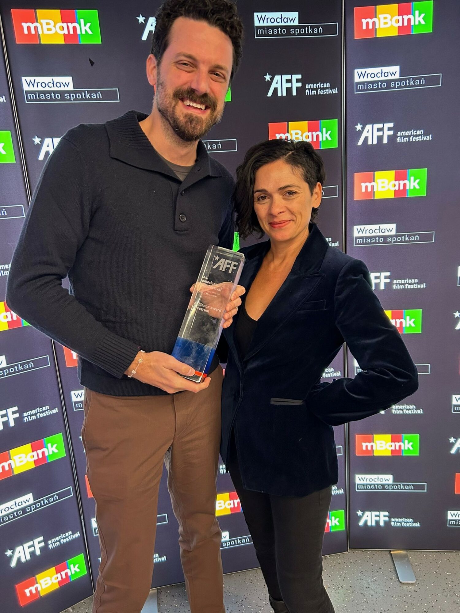 Man and woman standing together, holding an award, smiling, in front of a backdrop with logos and text.