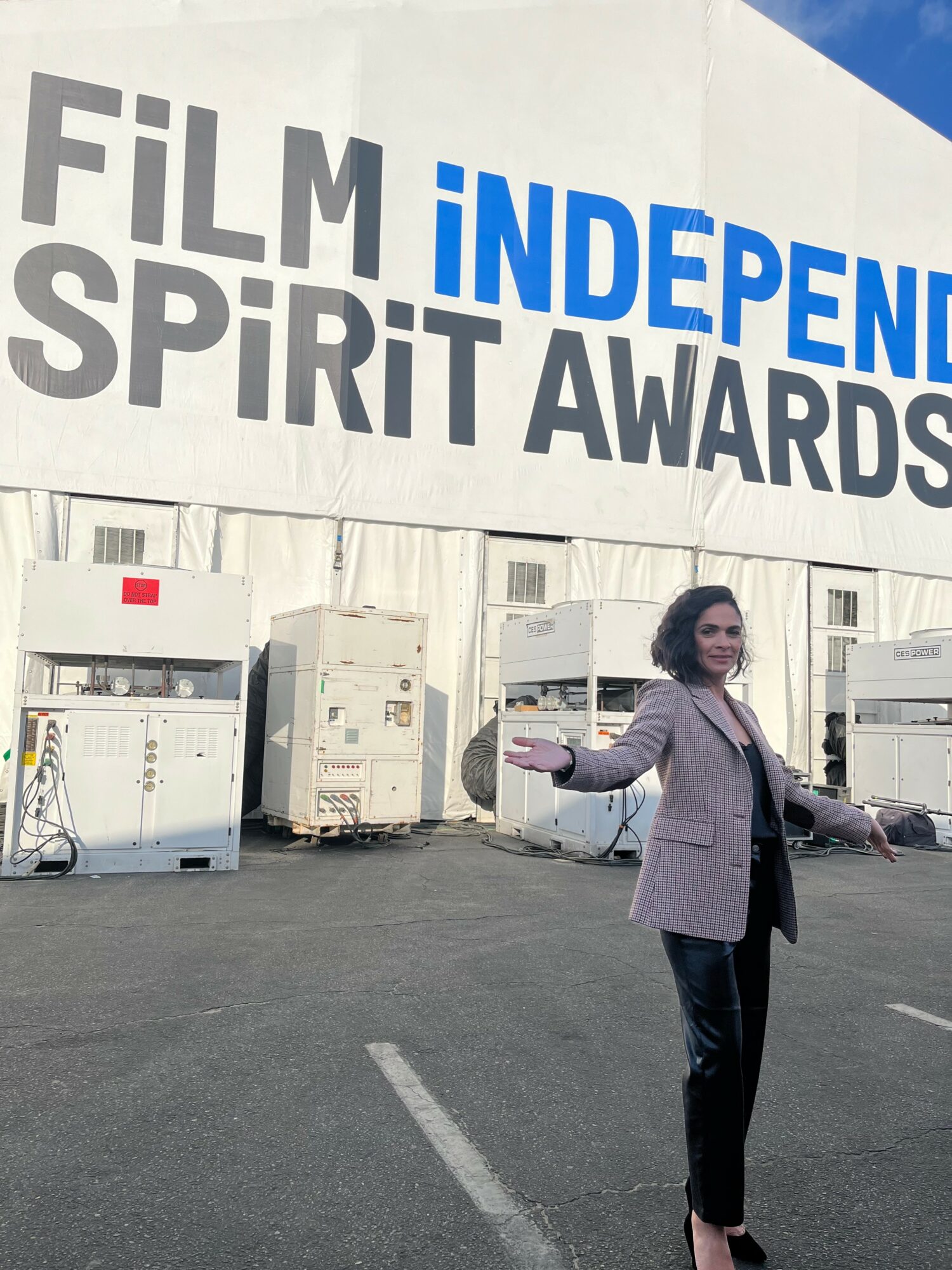 Woman standing in front of a large sign reading 'Film Independent Spirit Awards' outside, with equipment behind her.
