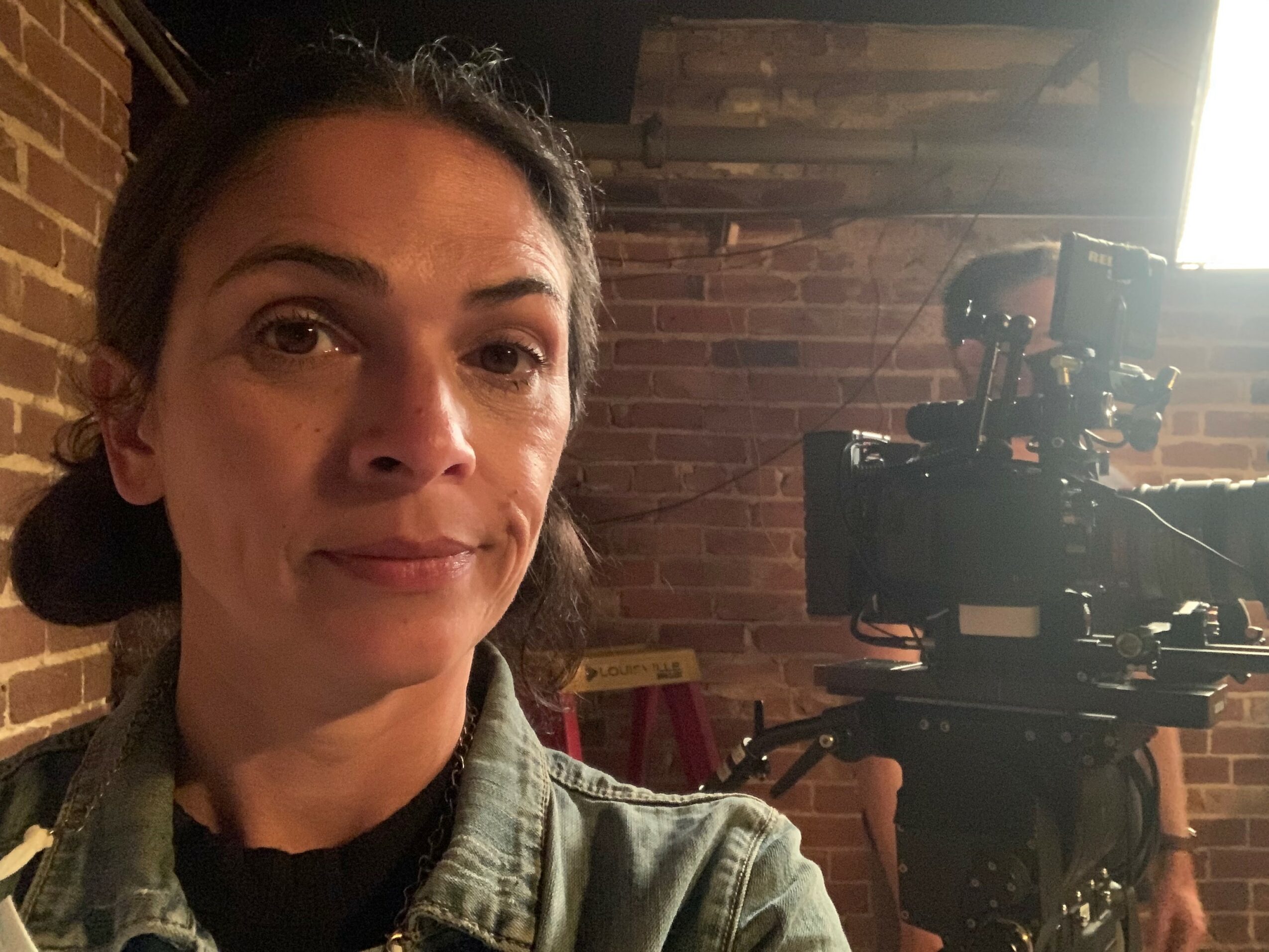 Woman with dark hair in a ponytail, wearing a green jacket, in a brick-walled room with camera equipment in the background.