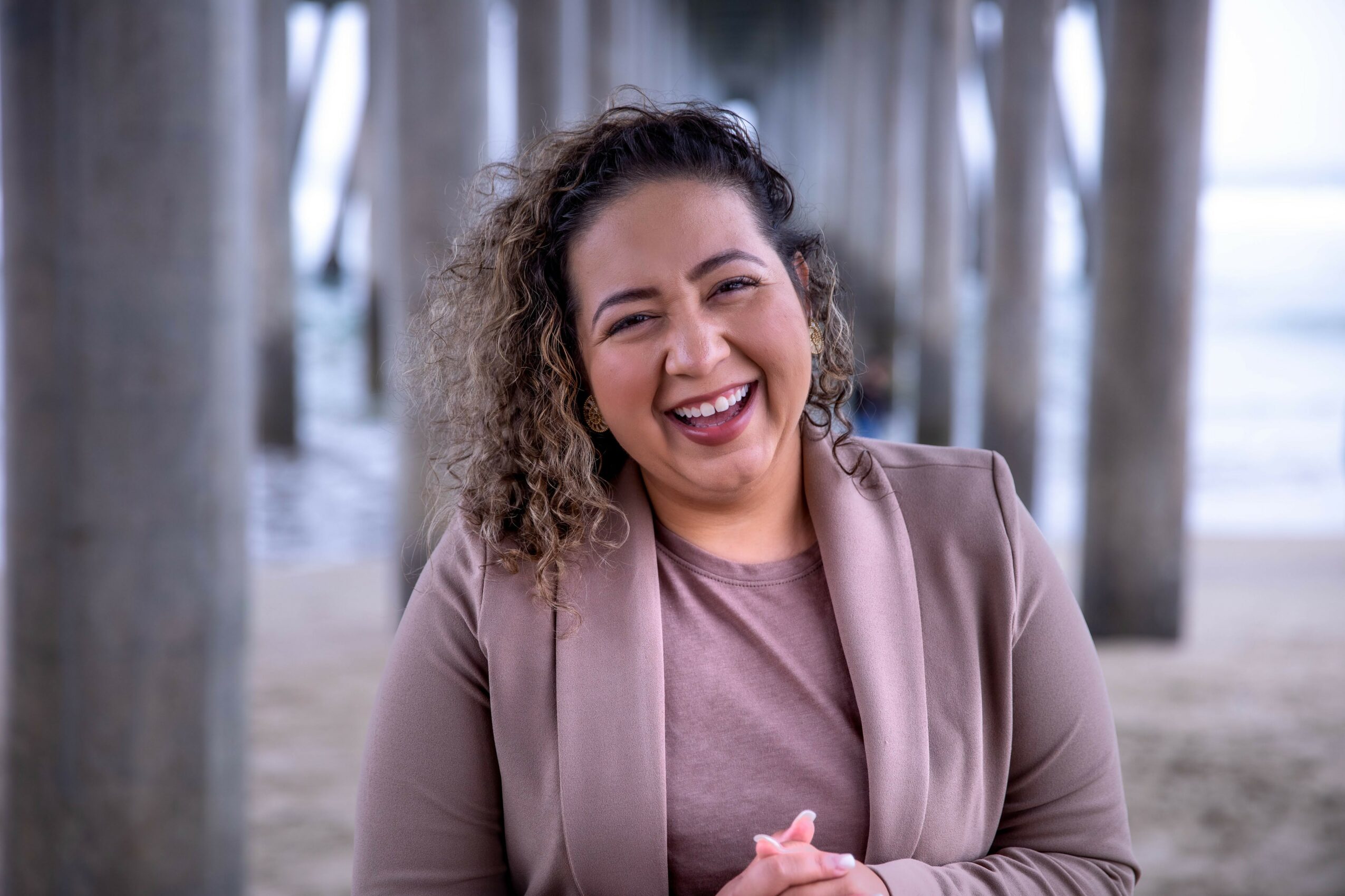 Smiling woman with curly hair under a pier, wearing a beige blazer and top, with a joyful expression.