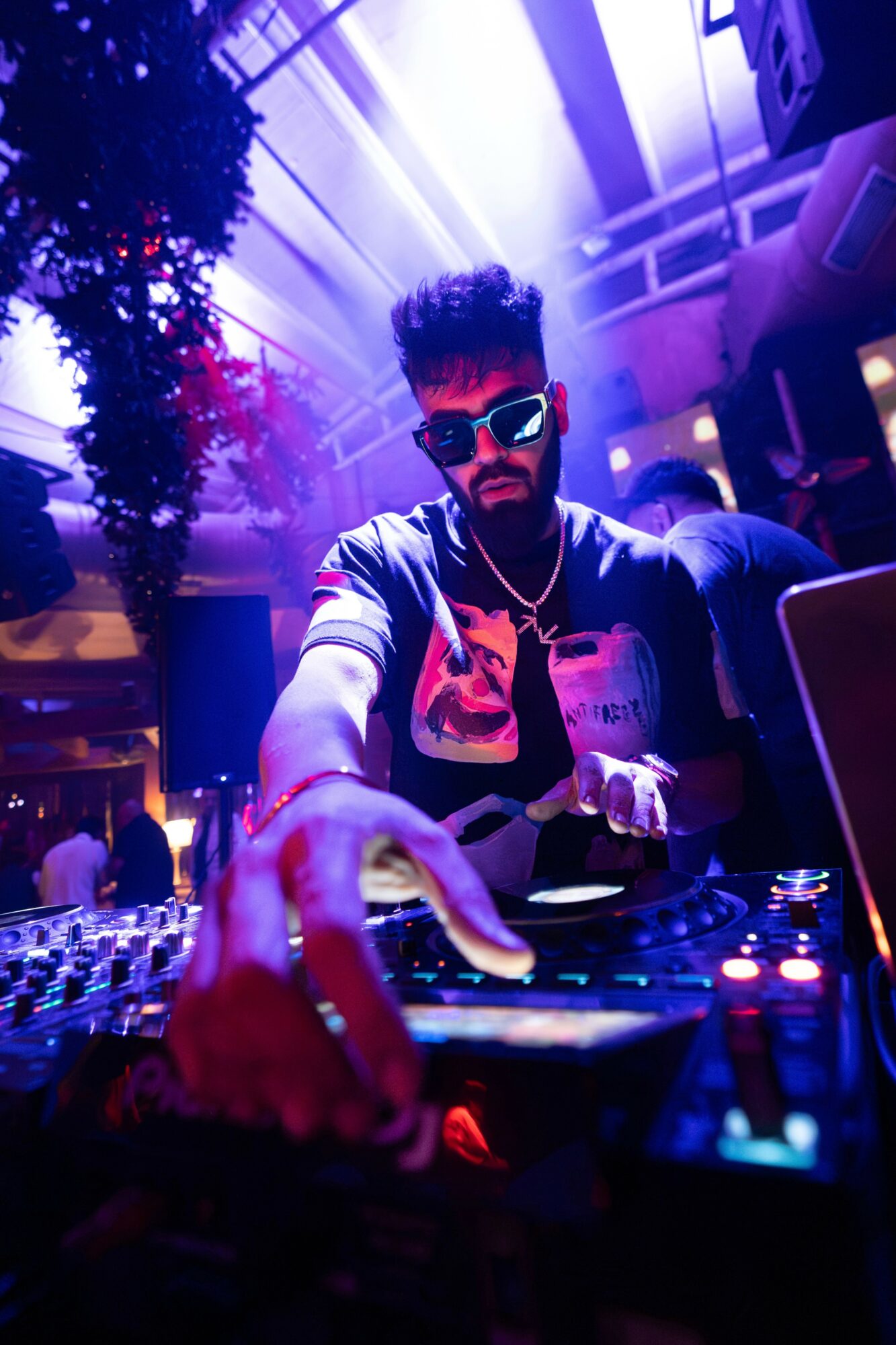 DJ with sunglasses and headphones performs at a nightclub with colorful lighting.