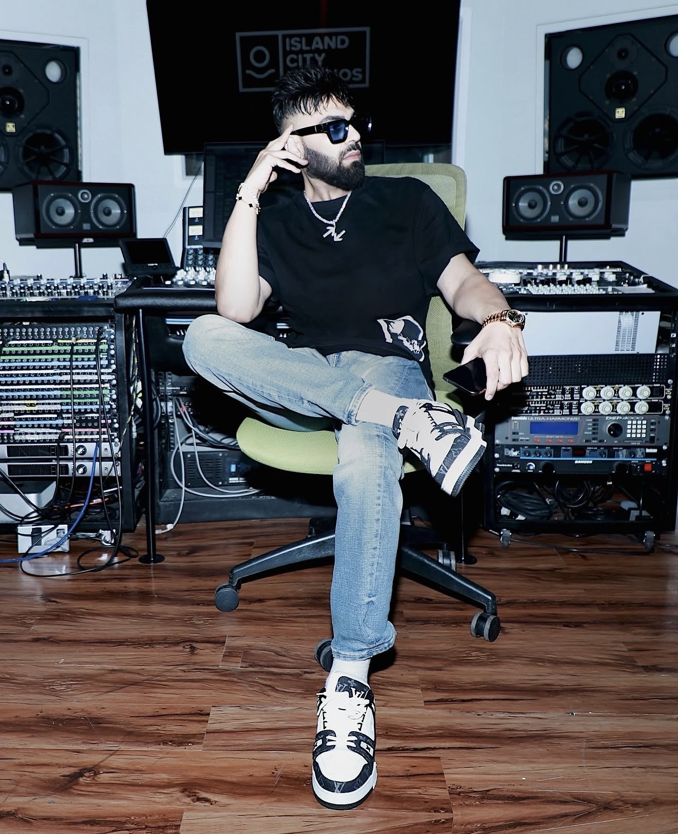 Person sitting in a music studio with audio equipment, wearing sunglasses, black t-shirt, jeans, and sneakers, with one hand on their head.