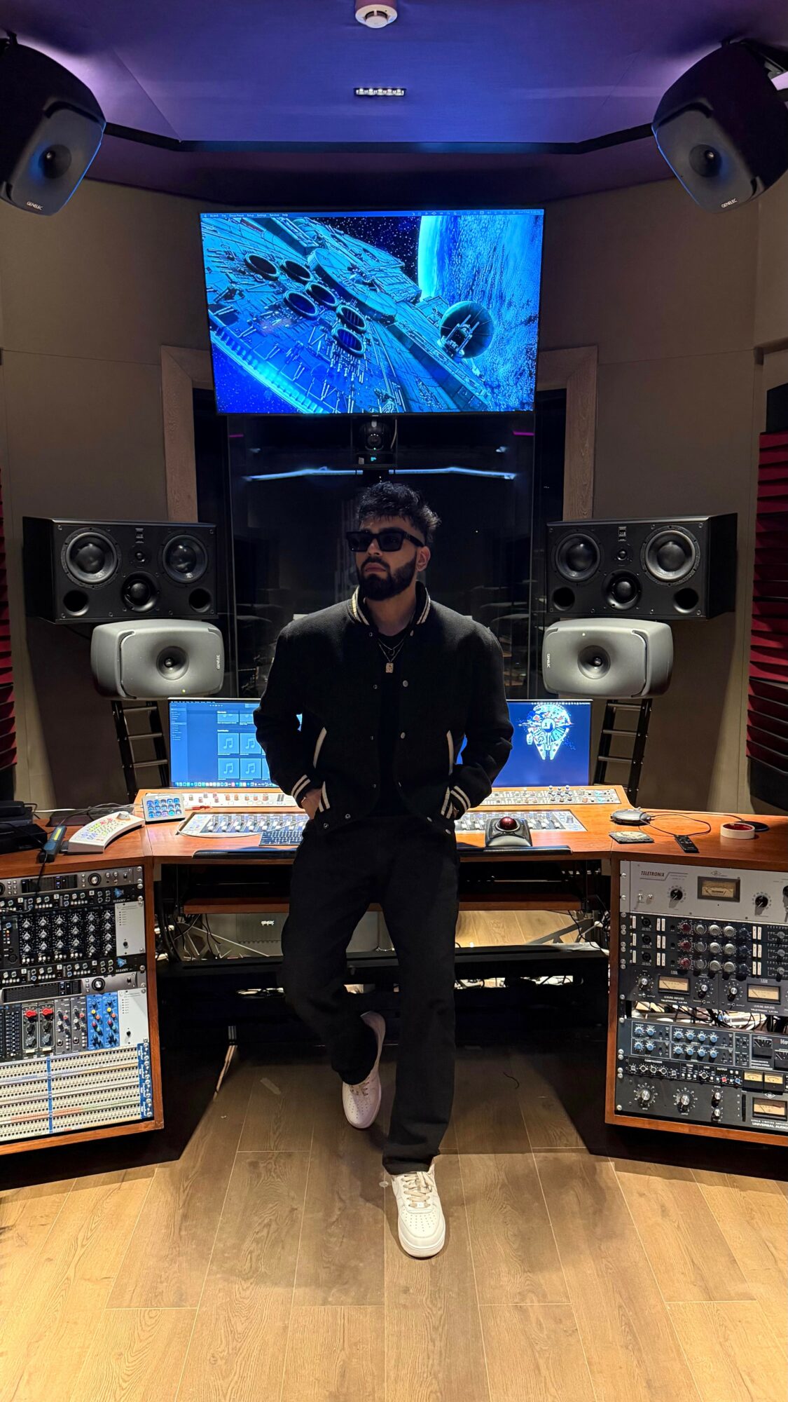 Man with sunglasses and beard standing in a music studio with equipment and large screens behind him.