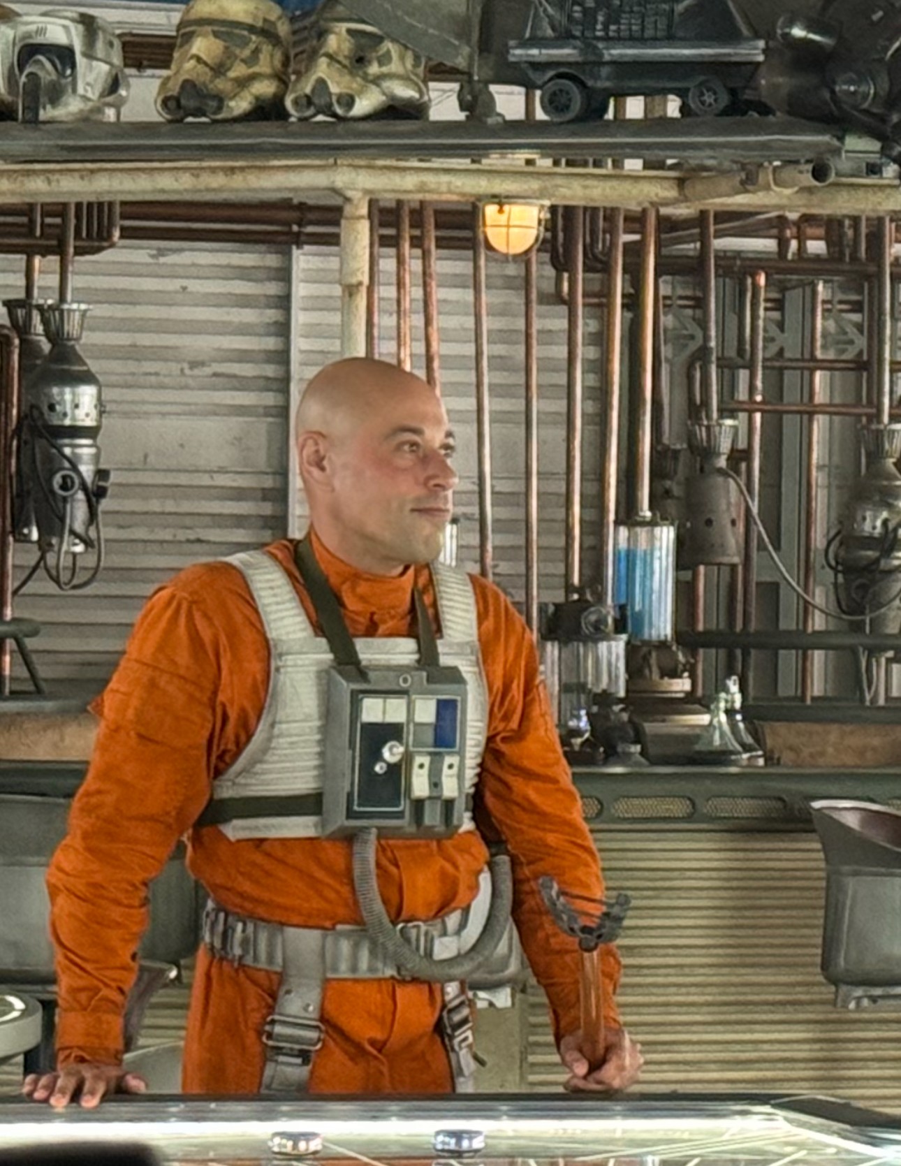 Person in orange jumpsuit with a vest, standing in a workshop with pipes and tools behind him.