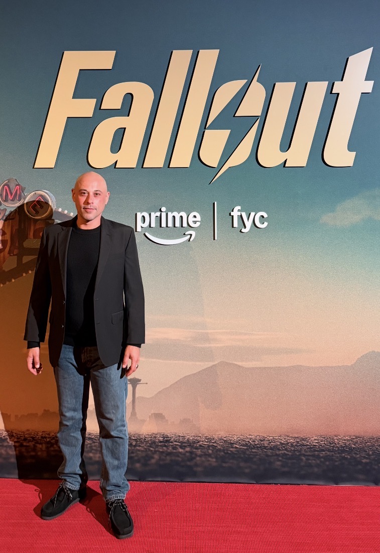 Person standing in front of a backdrop with the Fallout logo and a landscape scene, wearing a black blazer and jeans.