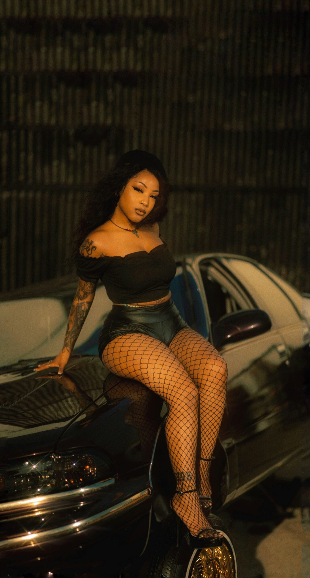Woman with long dark hair sitting on a car at night, wearing a black top, fishnet stockings, and high heels.