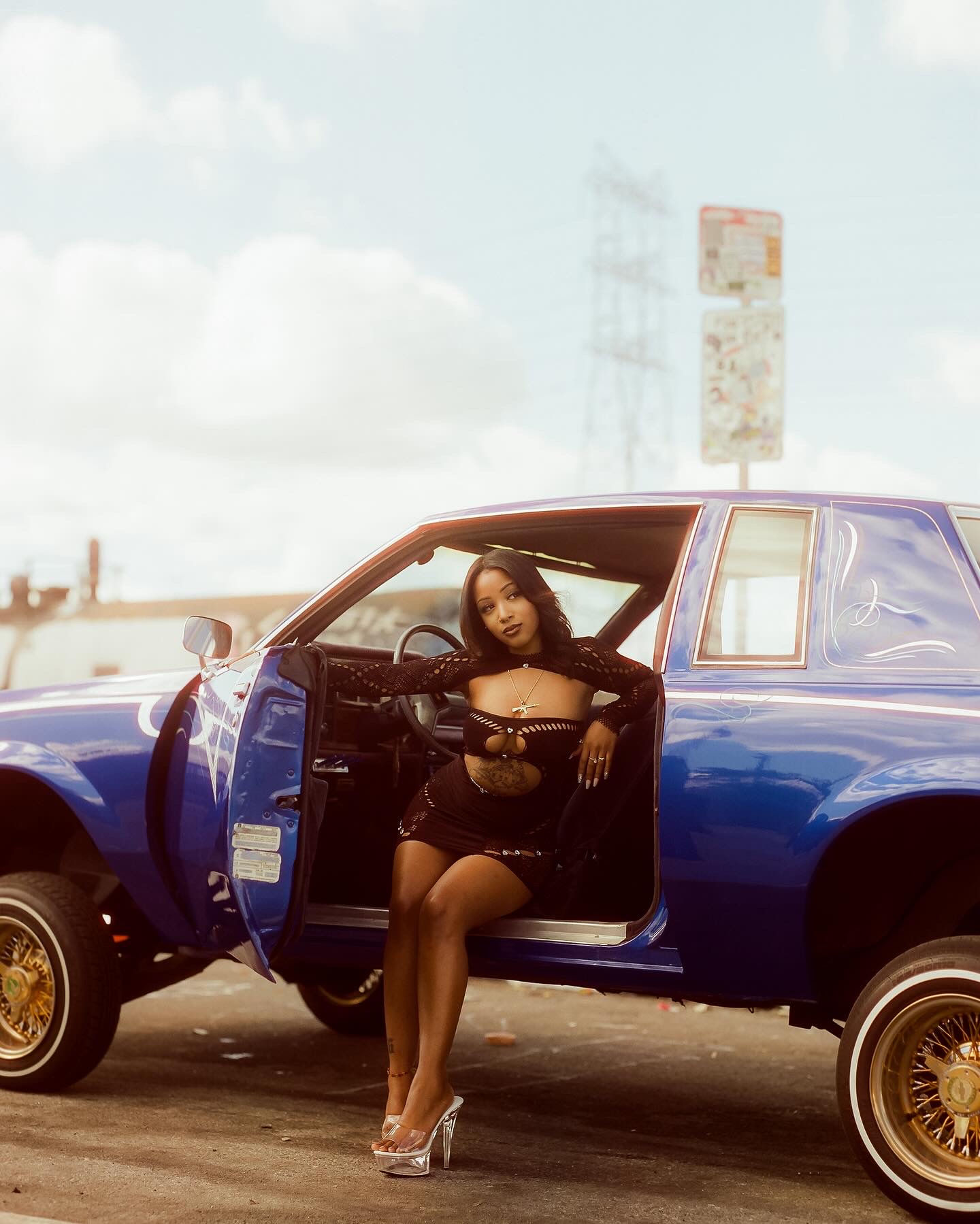 Woman sitting on car door with legs crossed, wearing black outfit, in an outdoor setting with a blue sky and clouds.