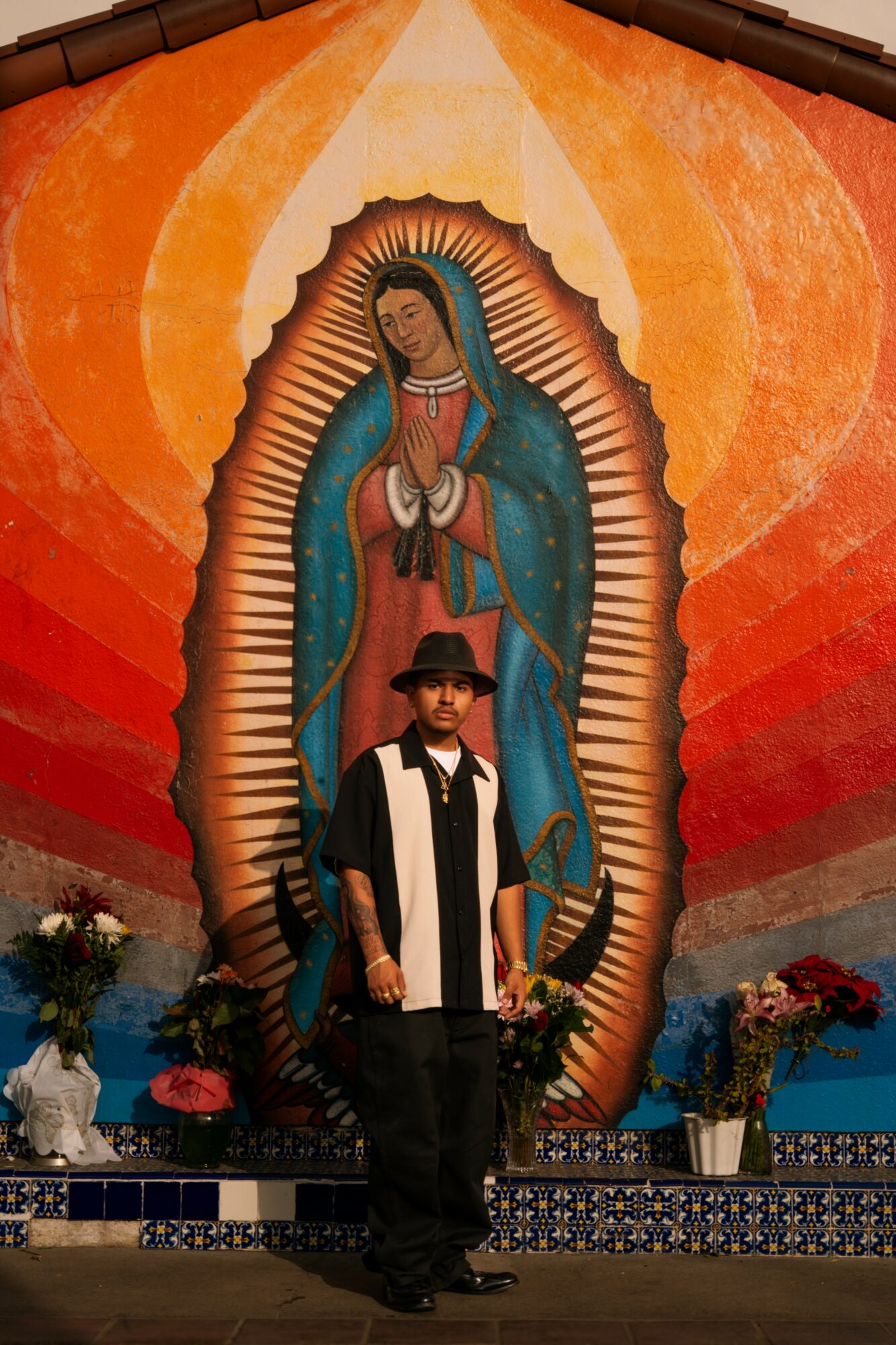 Person standing in front of a large religious image of the Virgin Mary with hands clasped, surrounded by flowers.
