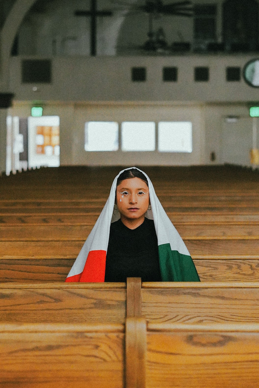 Woman with a headscarf sitting in a wooden pew inside a church, with a cross and windows in the background.
