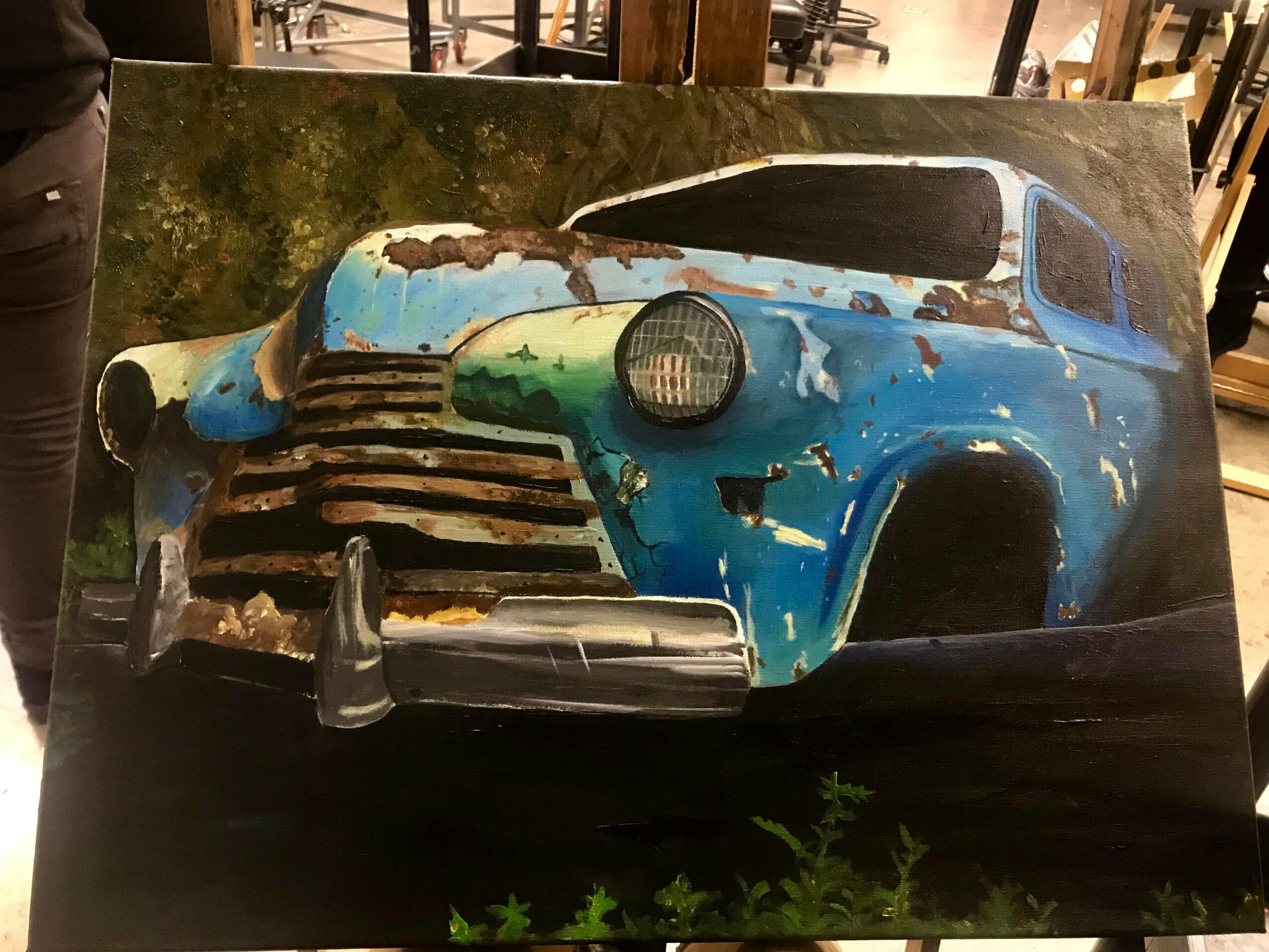 Rusty, abandoned blue car with missing parts and peeling paint, parked outdoors with greenery in the foreground.