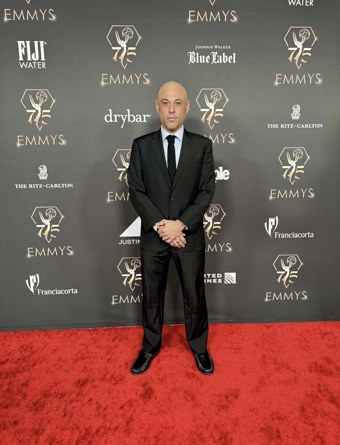 Man in black suit and tie standing on red carpet in front of a backdrop with logos and text.
