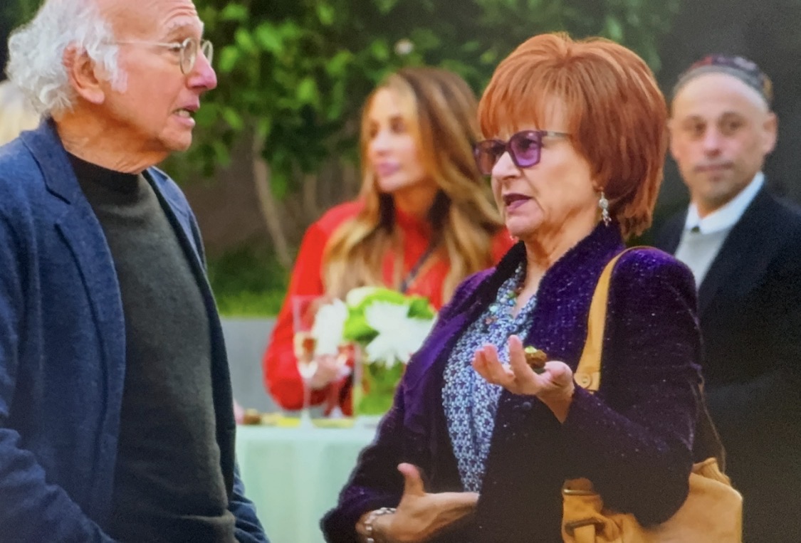 Four people outdoors, two in foreground talking, two in background, greenery, woman with glasses gesturing, man listening.