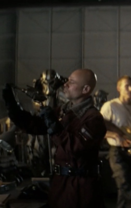 Bald man in uniform holding a camera, with another person in the background, in an indoor setting.
