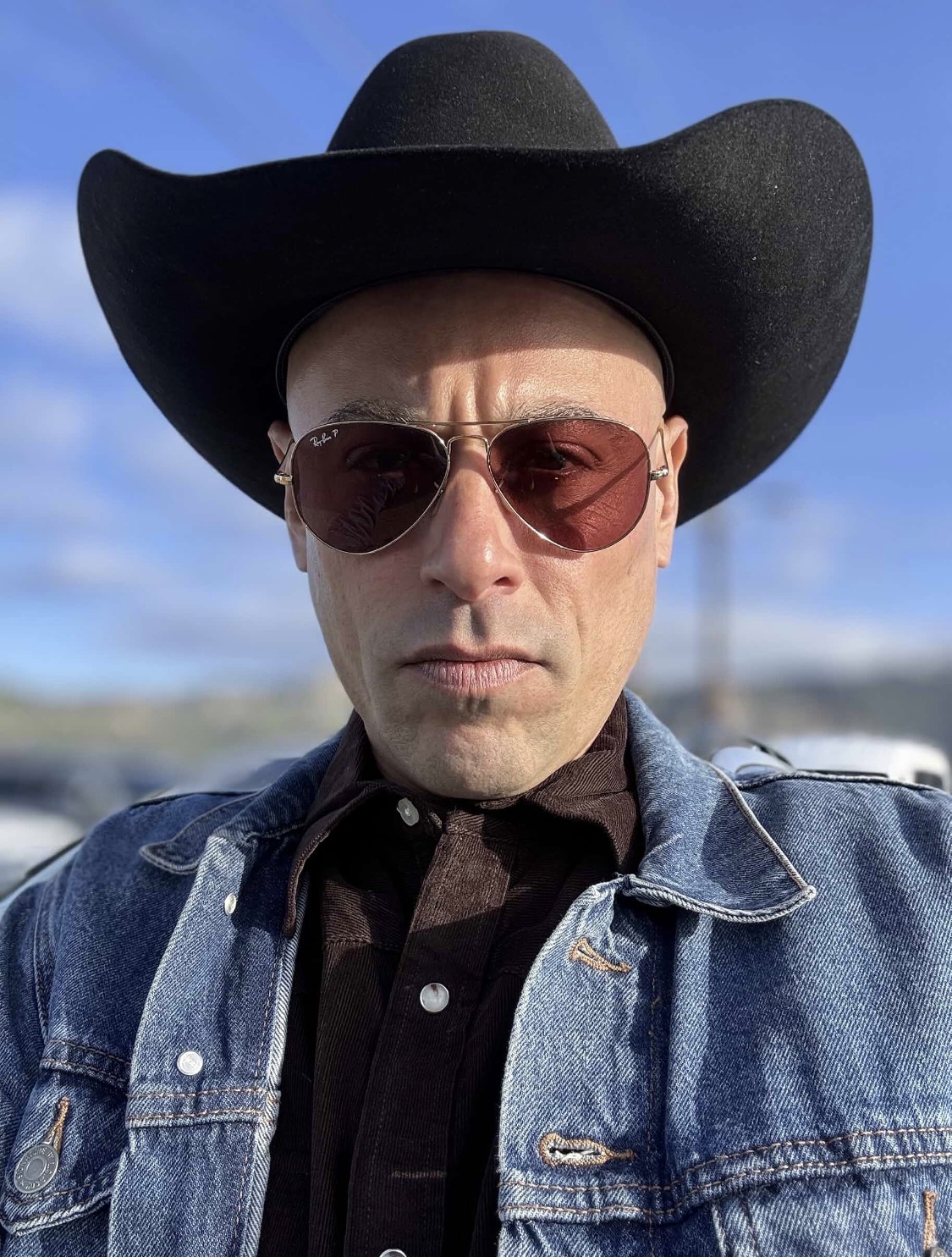 Person wearing a large black hat, sunglasses, denim jacket, and black shirt outdoors with a blue sky background.