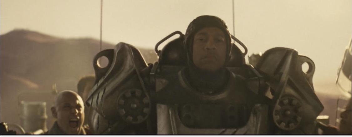 Person in a futuristic suit with large shoulder armor, outdoors with mountains in the background.