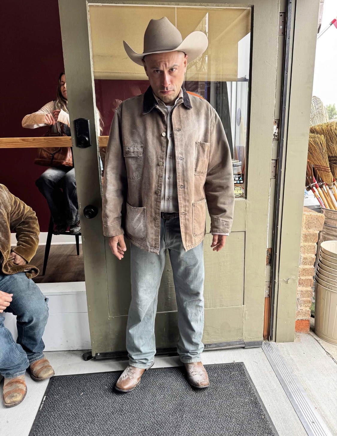 Man wearing a cowboy hat, jacket, and jeans standing inside a glass booth, with a person sitting nearby in a cafe.