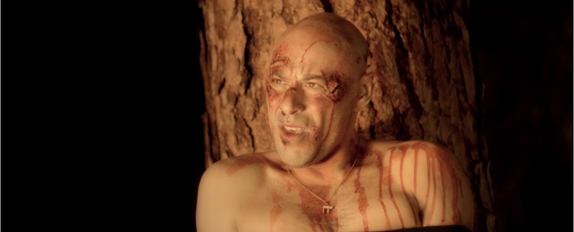 Man with a shaved head and beard, shirtless, leaning against a tree, with blood on his chest and face.