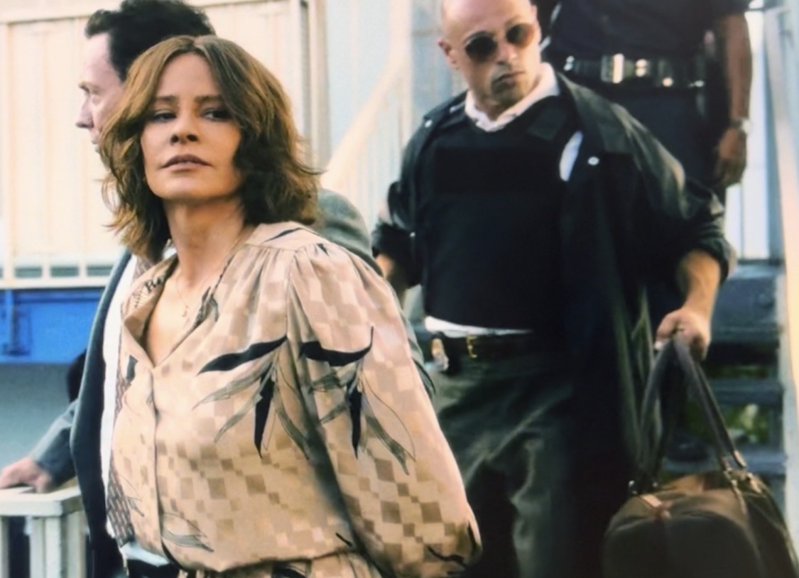 Woman with shoulder-length hair in patterned blouse walking outdoors, man with sunglasses and backpack behind her, carrying a bag.