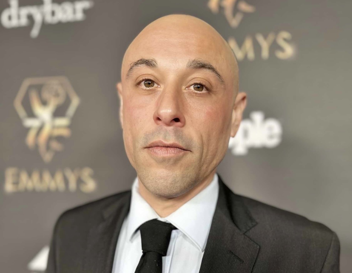 Bald man in a suit and tie at an awards event, background with logos and text