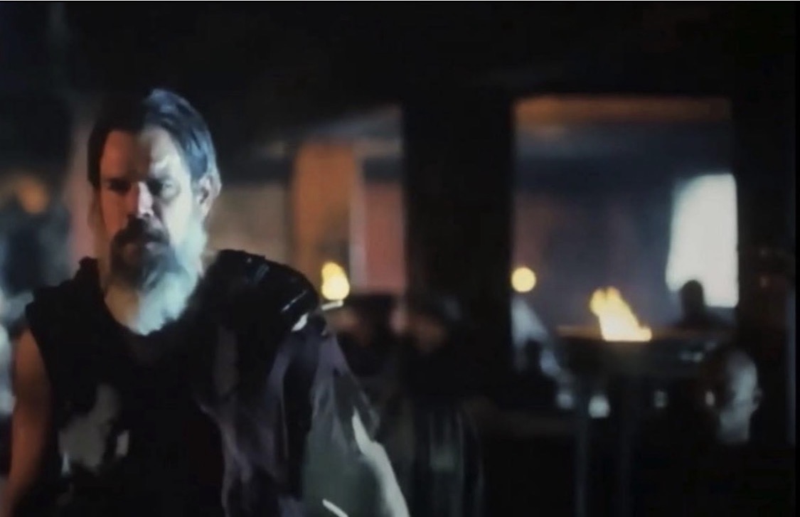 A man with a beard and dark clothing in a dimly lit indoor setting with fire and cars in the background.